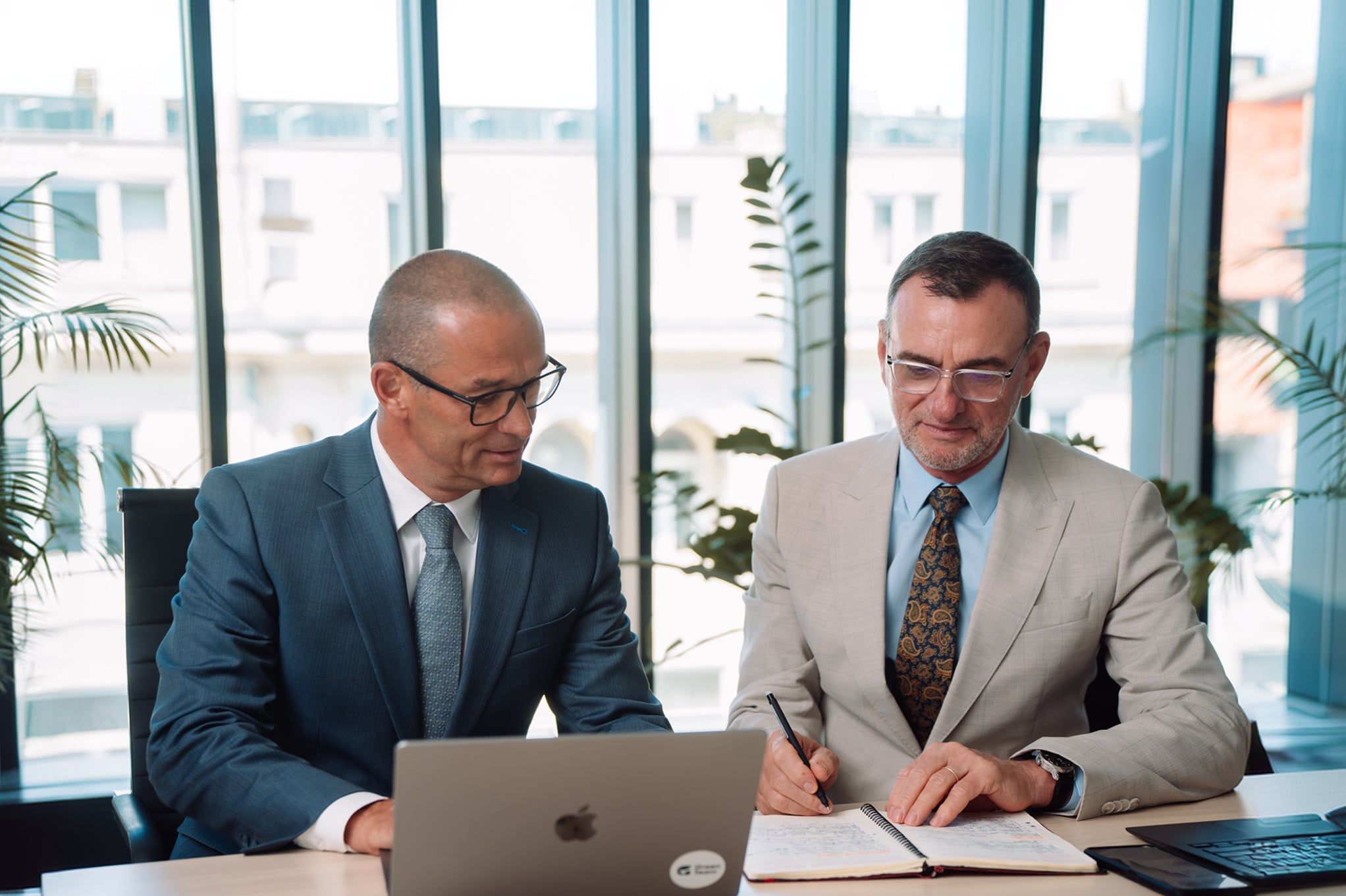Two senior advisors at a meeting table, one taking notes beside an open laptop.