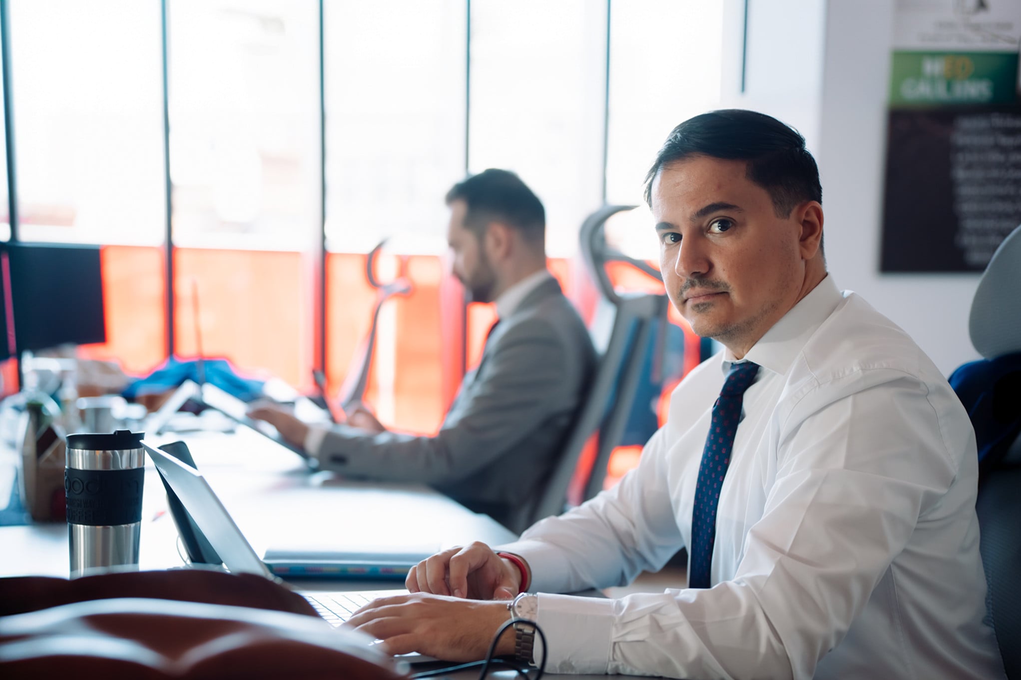 Advisor working on a laptop in an open-plan office, with a colleague in the background.